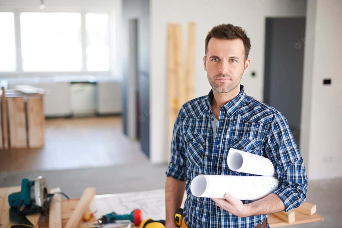 travailleur du batiment en chemise a carreaux avec un mètre mesureur accroché à la ceinture et deux plans sous le bras