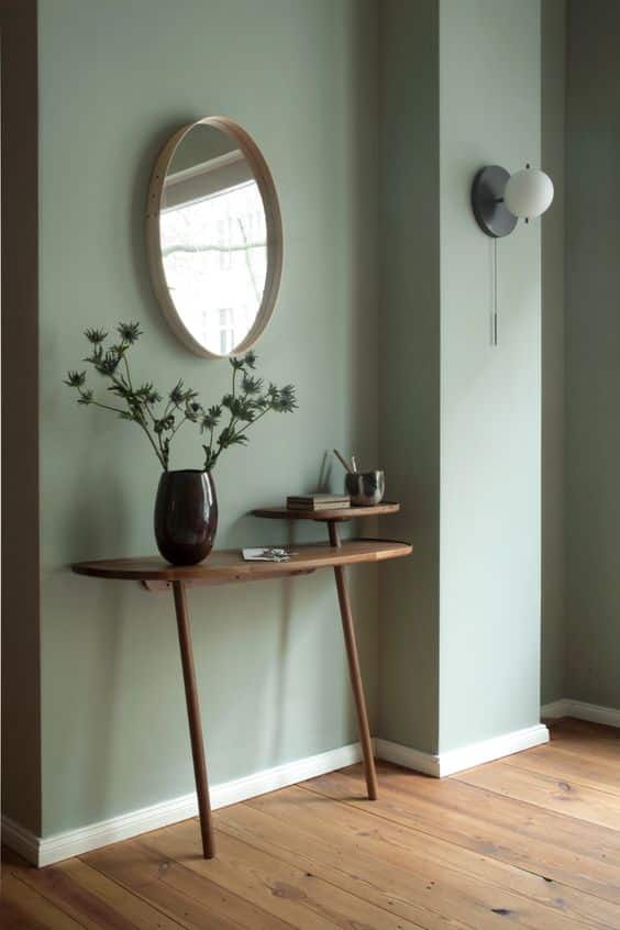 Une entrée avec un miroir rond un meuble en noyer et un mur vert céladon