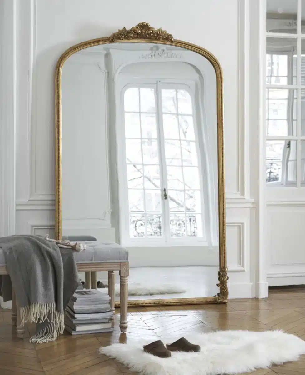 Un grand miroir avec moulure et cadre doré posé au sol dans un salon avec mur blanc et parquet chevron