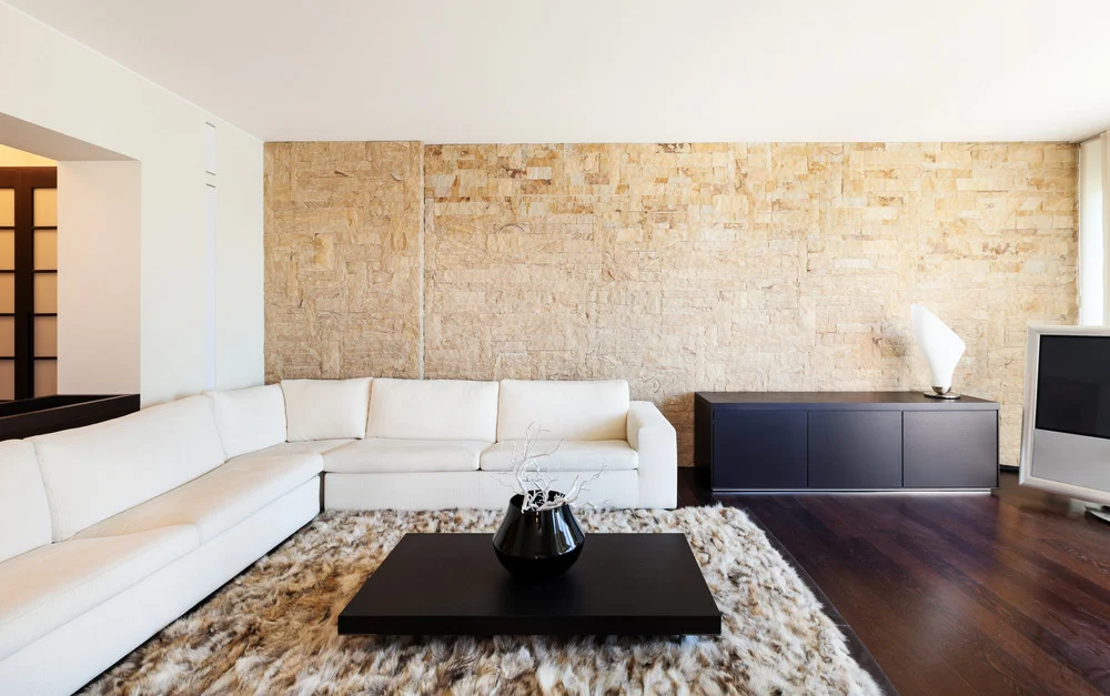 Salon contemporain avec un mur en pierre claire derrière un grand canapé d’angle blanc